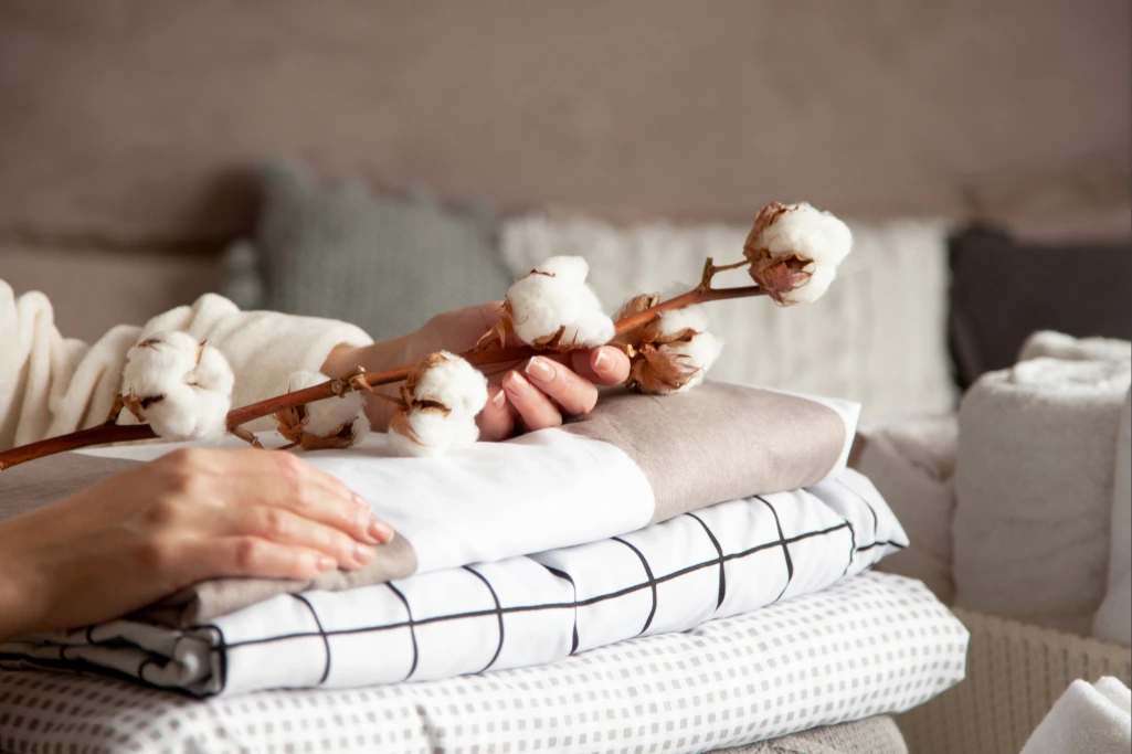 Mãos femininas repousam sobre uma pilha de tecidos bem dobrados, segurando delicadamente um ramo de algodão. A imagem conecta a matéria-prima natural ao produto final, simbolizando o conforto e a qualidade do algodão brasileiro.