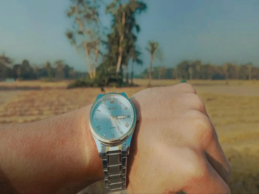 Close de um relógio de pulso elegante usado na mão de um homem, capturado à luz do dia natural com um campo rural e árvores ao fundo.