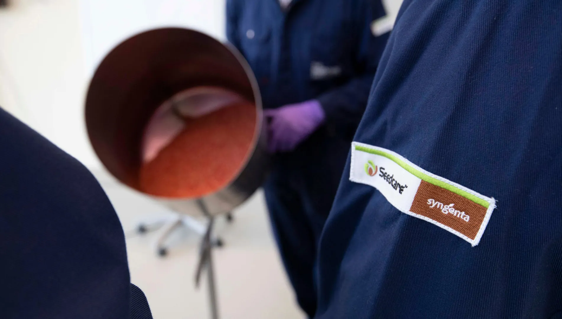 profissionais da Syngenta Seedcare em ambiente de tratamento de sementes, com equipamento ao fundo e detalhe do uniforme com logomarca da marca