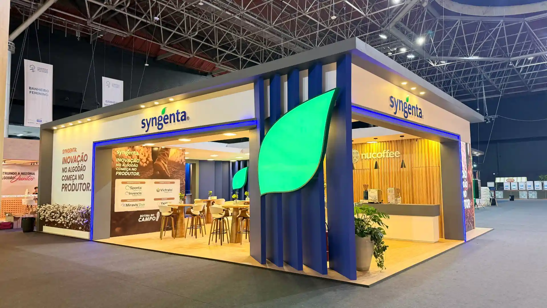Estande corporativo da Syngenta no 15º Congresso Brasileiro do Algodão o que marcou o evento , caracterizado por uma grande estrutura moderna com acabamento em tons de cinza, branco, azul e detalhes em madeira. Na fachada e nas laterais destacam-se o logotipo da marca com uma folha verde, além de um painel retroiluminado com o texto "INOVAÇÃO NO ALGODÃO COMEÇA NO PRODUTOR" e marcas de produtos como Sponta, Invencis, Victrato, Miravis Duo e YieldOn. 