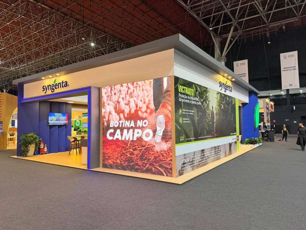 Estande corporativo da Syngenta no 15º Congresso Brasileiro do Algodão, exibindo uma arquitetura moderna com acabamentos em tons de cinza, branco e azul, além de piso em tom amarelado. Na fachada principal, destaca-se o logotipo da marca acompanhado por um grande painel luminoso vertical com a frase "BOTINA NO CAMPO" sobre uma imagem de solo agrícola e pés com botas. Na lateral direita, o estande apresenta um banner publicitário de Victrato com textos descritivos e avisos legais, enquanto no interior vislumbrar-se mesas de atendimento e uma televisão fixada na parede. O teto do pavilhão exibe uma ampla estrutura metálica treliçada industrial.