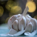 Cabeça de alho com dentes à mostra e cascas soltas, sobre superfície azulada com luzes desfocadas ao fundo