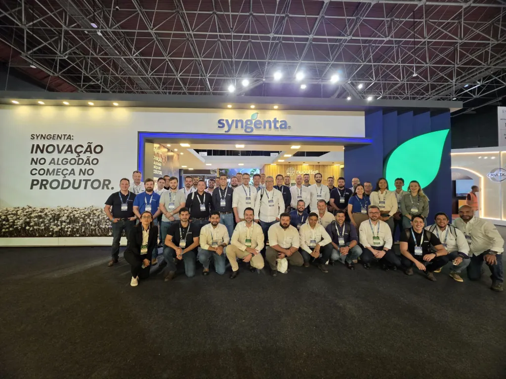 Fotografia de grupo com a equipa da Syngenta posando em frente ao seu estande no 15º Congresso Brasileiro do Algodão. Os colaboradores, vestindo crachás e roupas corporativas ou casuais, encontram-se dispostos em fileiras (sentados à frente e em pé atrás) no piso escuro de um pavilhão de feiras. À esquerda, vê-se um painel branco com o logotipo da marca e a frase "SYNGENTA: INOVAÇÃO NO ALGODÃO COMEÇA NO PRODUTOR.", além de uma decoração simulando capulho de algodão. Ao fundo, ergue-se a estrutura do estande com a logomarca iluminada, detalhes em azul e uma imensa folha verde decorativa, sob o teto metálico industrial do pavilhão.   